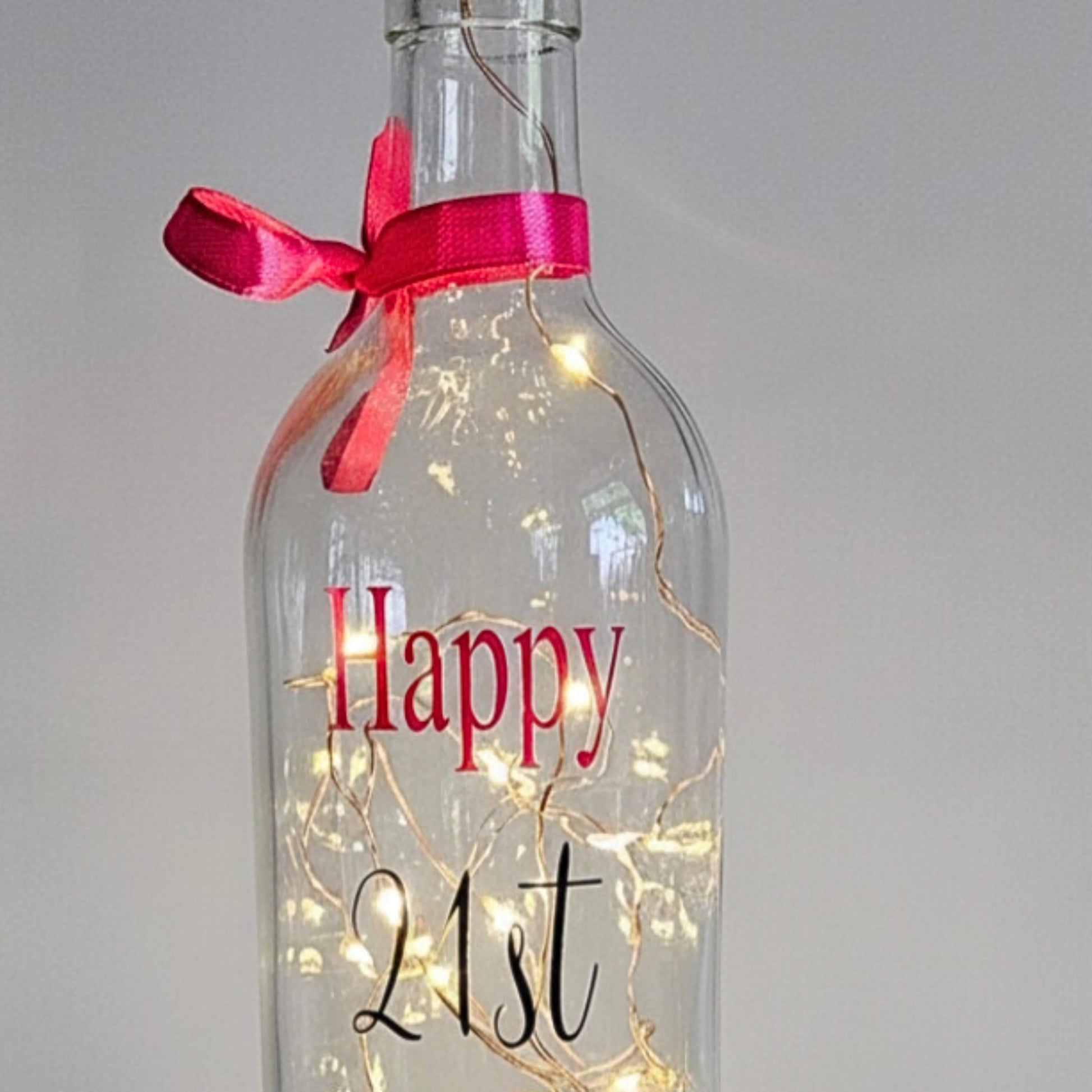 A clear bottle with lights. The word 'Happy' in pink & 21st in black. Has a pink ribbon around the neck
