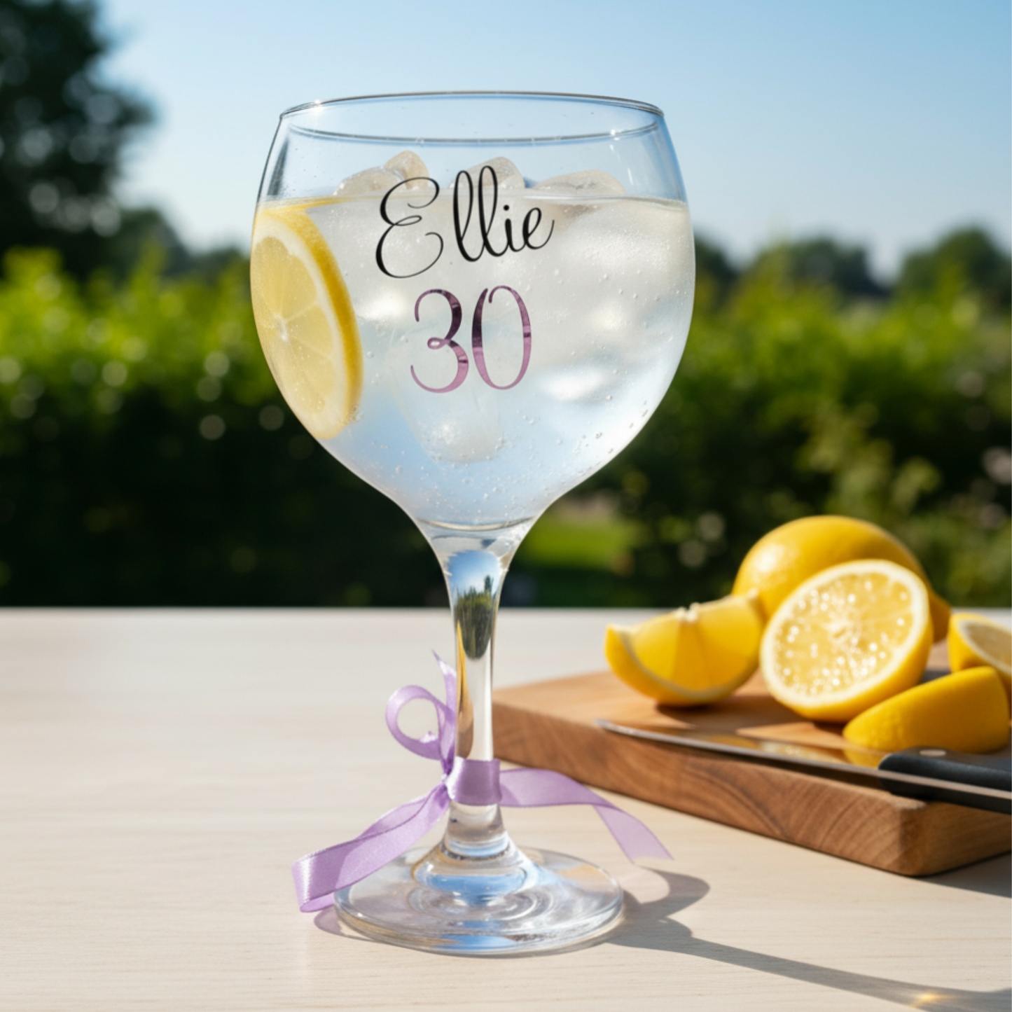 Personalised gin & cocktail glass with the name 'Ellie' in glossy black vinyl and age '30' in purple pink holographic vinyl. Sat on a white surface next to a chopping board of lemons in a garden setting.