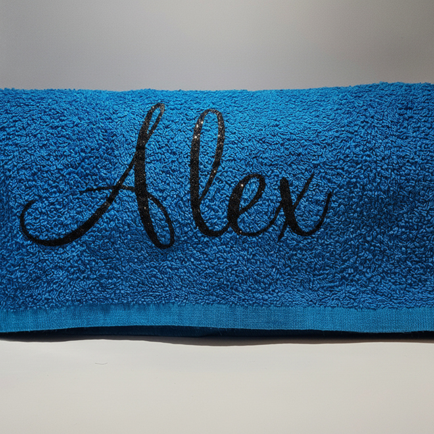 Personalised royal blue beach towel with the name 'Alex' in a black glitter vinyl on a neutral background