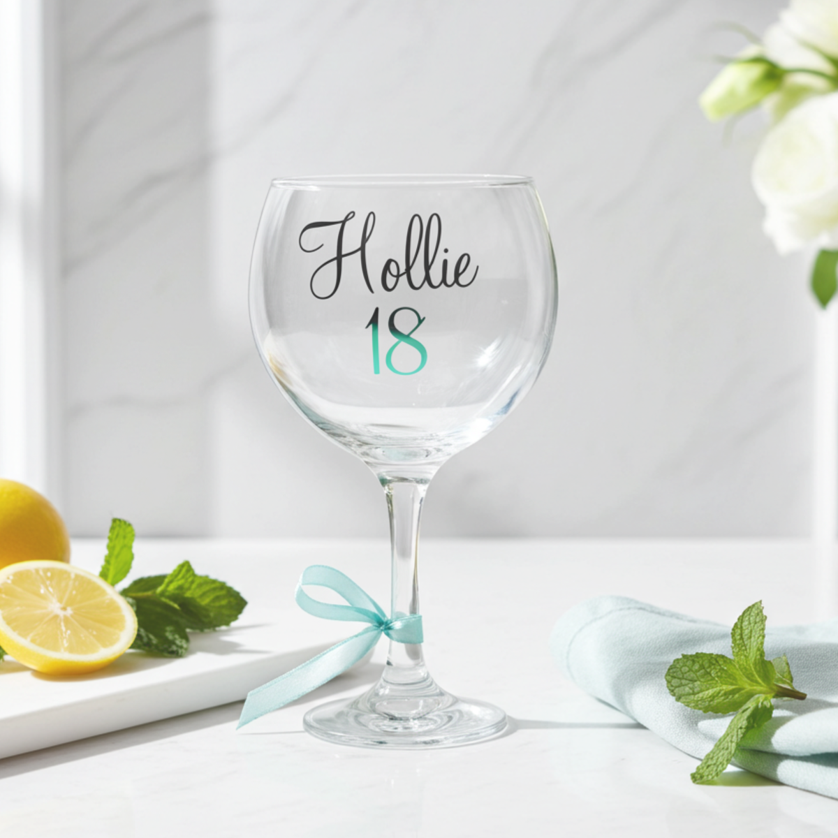 Personalised balloon gin glass with the name 'Hollie' in a flowing script font from a glossy black vinyl. This stemmed cocktail goblet features age '18' in a mint chrome holographic vinyl & a mint satin ribbon tied around its stem. Placed on a white marble surface next to a chopping board of lemon slices & mint sprigs.