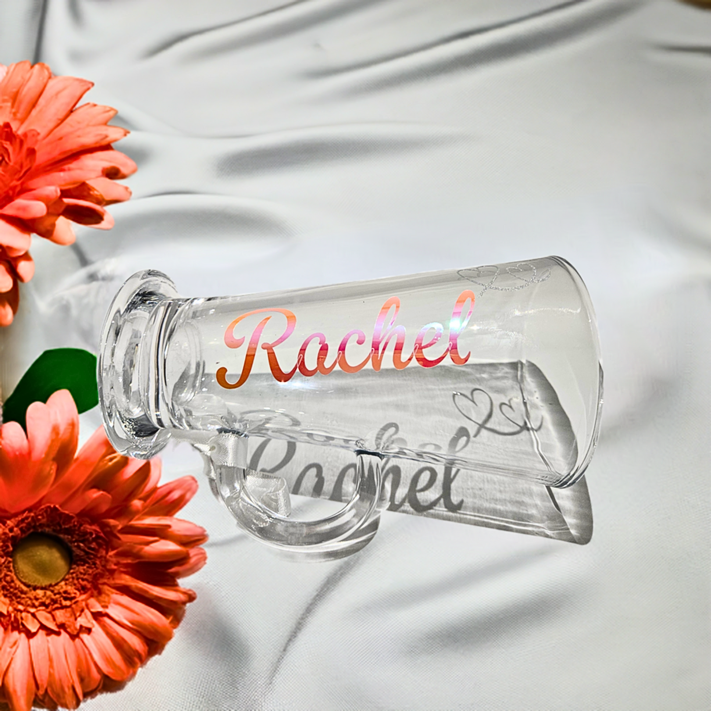 Clear glass mug with 'Rachel' in a holographic vinyl & silver glitter heart. Staged with orange flowers on a satin drape