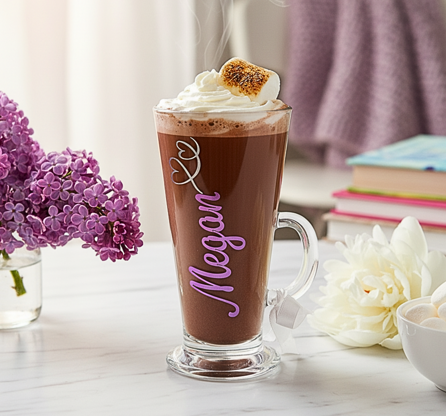 Hot chocolate in a glass mug with whipped cream and a marshmallow, placed on a table with flowers and books.