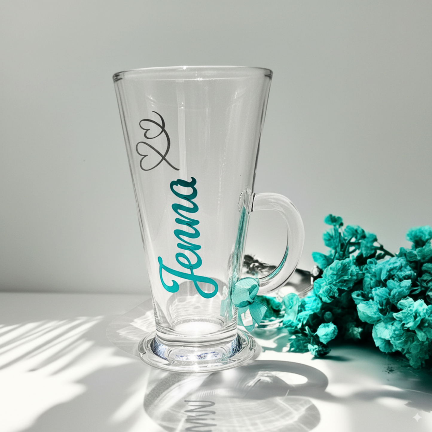 Custom name personalised clear glass hot chocolate mug with 'Jenna' on it in a premium mint chrome vinyl with delicate swirl heart decoration in silver glitter vinyl. Presented with a luxury mint coloured satin ribbon on the handle.