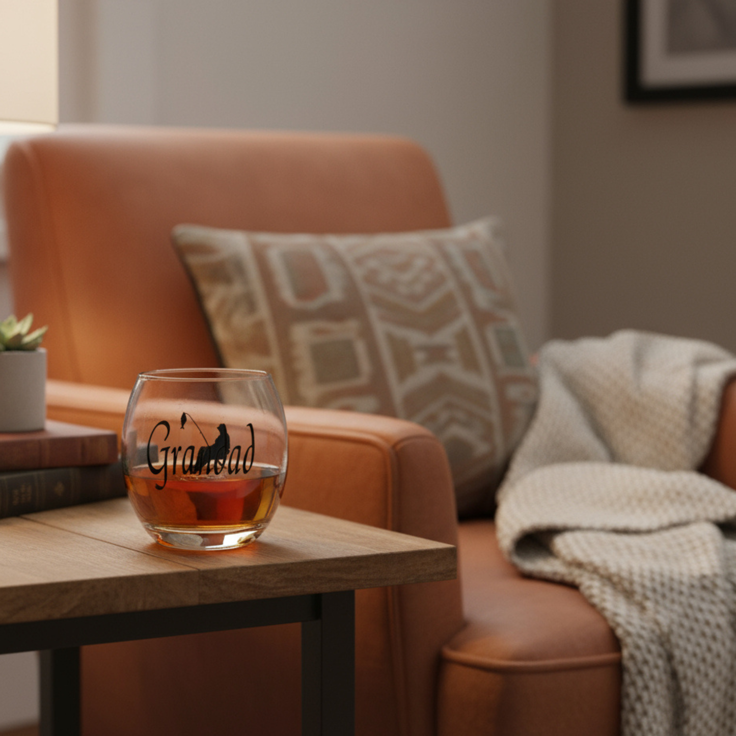 Personalised whisky glass tumbler with 'Grandad' on the front. Sat on a wooden table next to an orange armchair with cream knit blanket & cushion.