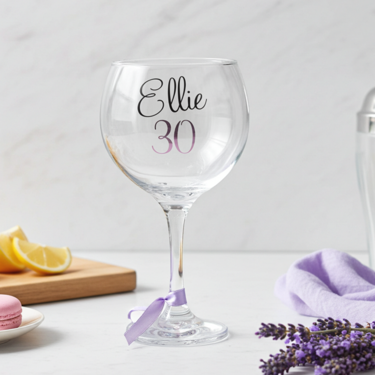Personalised balloon gin glass with the name 'Ellie' in a flowing script font designed from a glossy black vinyl. This cocktail goblet has age '30' in a pink purple chrome holographic vinyl and a lavender satin ribbon around its stem. Styled on a white surface next to lavender sprigs & a chopping board of sliced lemons.