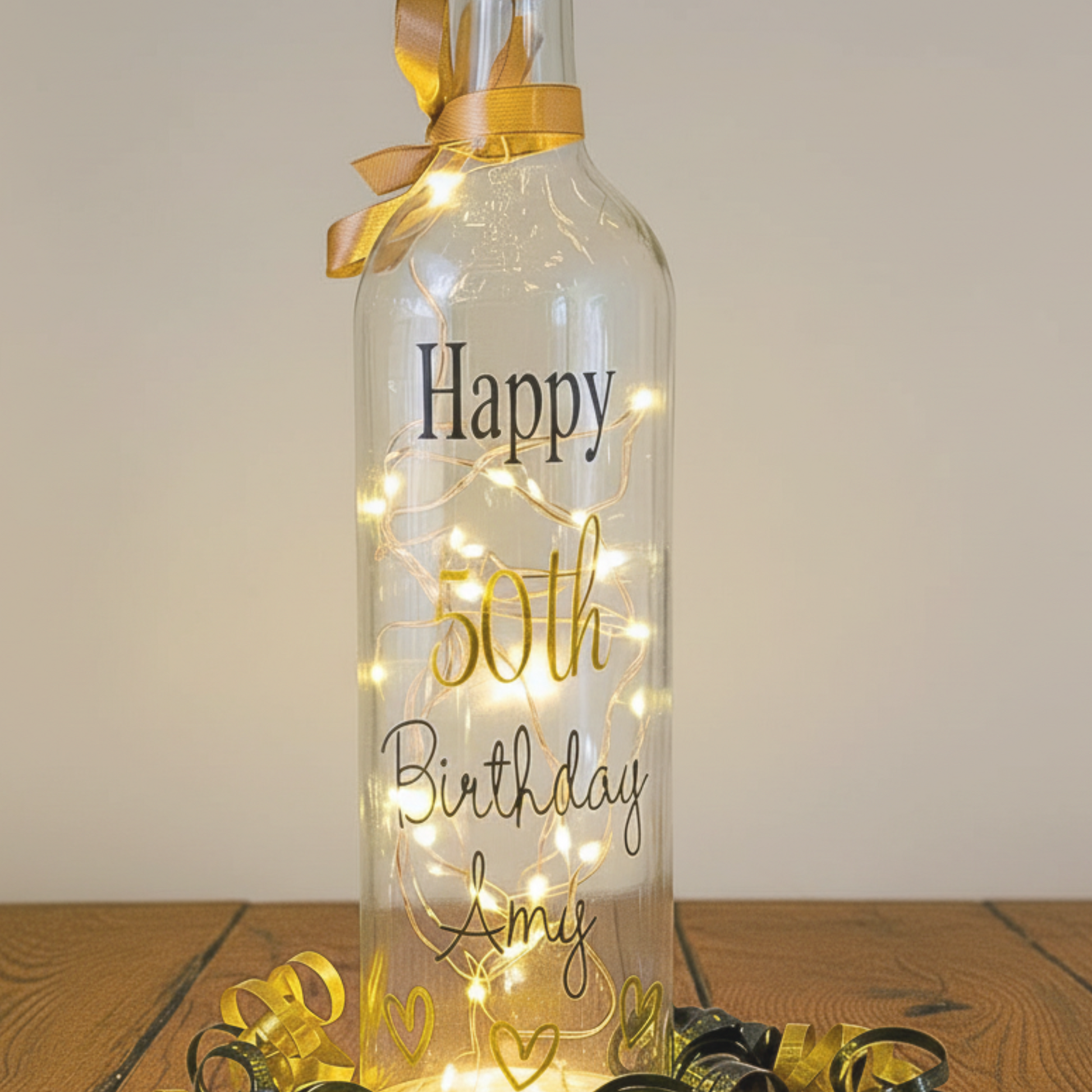 Personalised 50th LED light up glass bottle with 'Happy 50th Birthday Amy' text in glossy gold & black vinyl on a wooden surface with satin curling ribbons around.