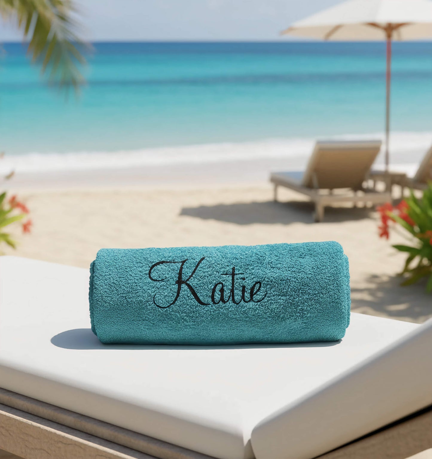 Teal towel with 'Katie' in a black glitter vinyl on it, placed on a lounge chair by the beach.