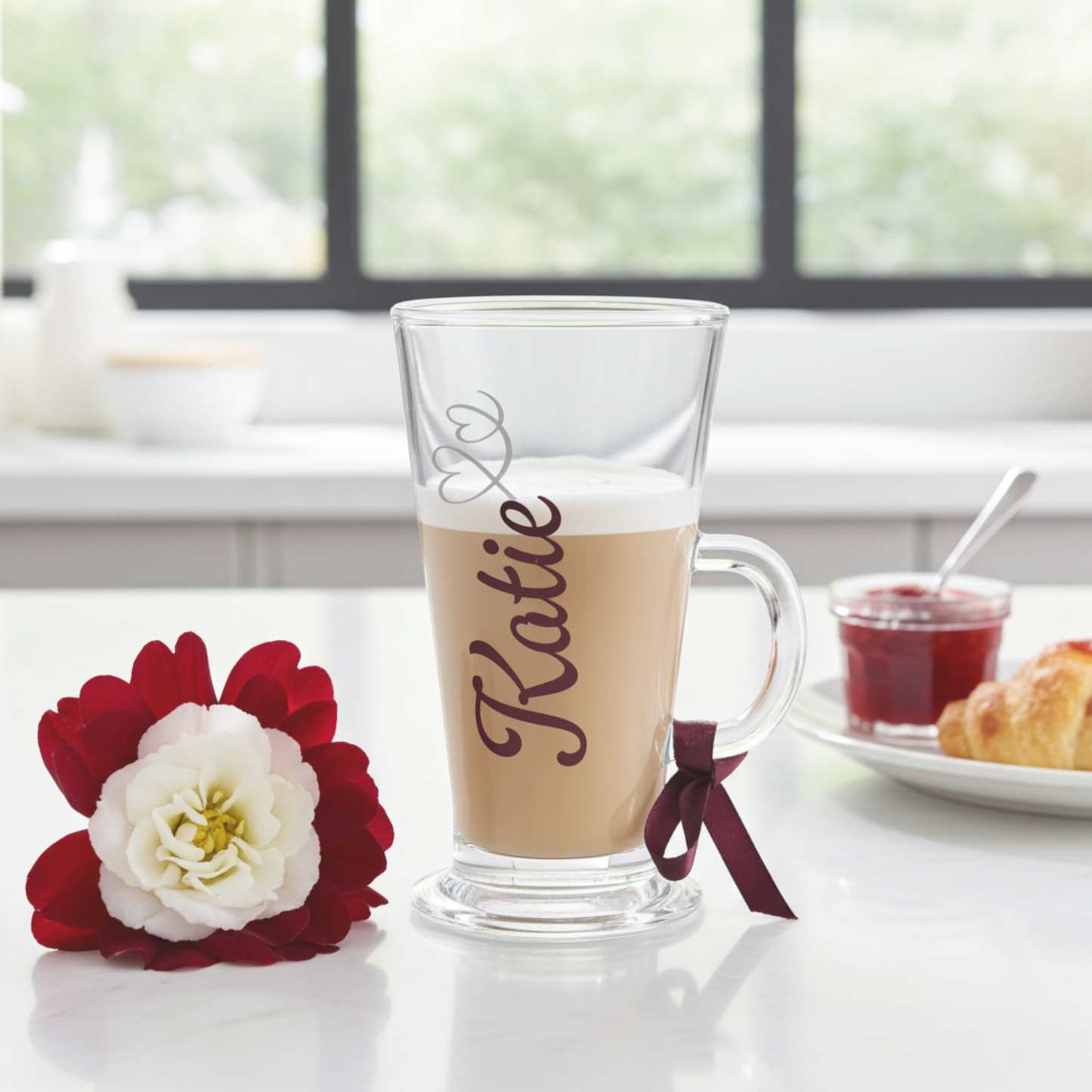 Personalised latte glass mug with the text 'Katie' on it in a holographic chrome burgundy vinyl. Features a silver glitter heart swirl design & a burgundy satin ribbon on the handle. Placed on a marble surface next to a burgundy flower & croissant with a jam pot on a small plate.