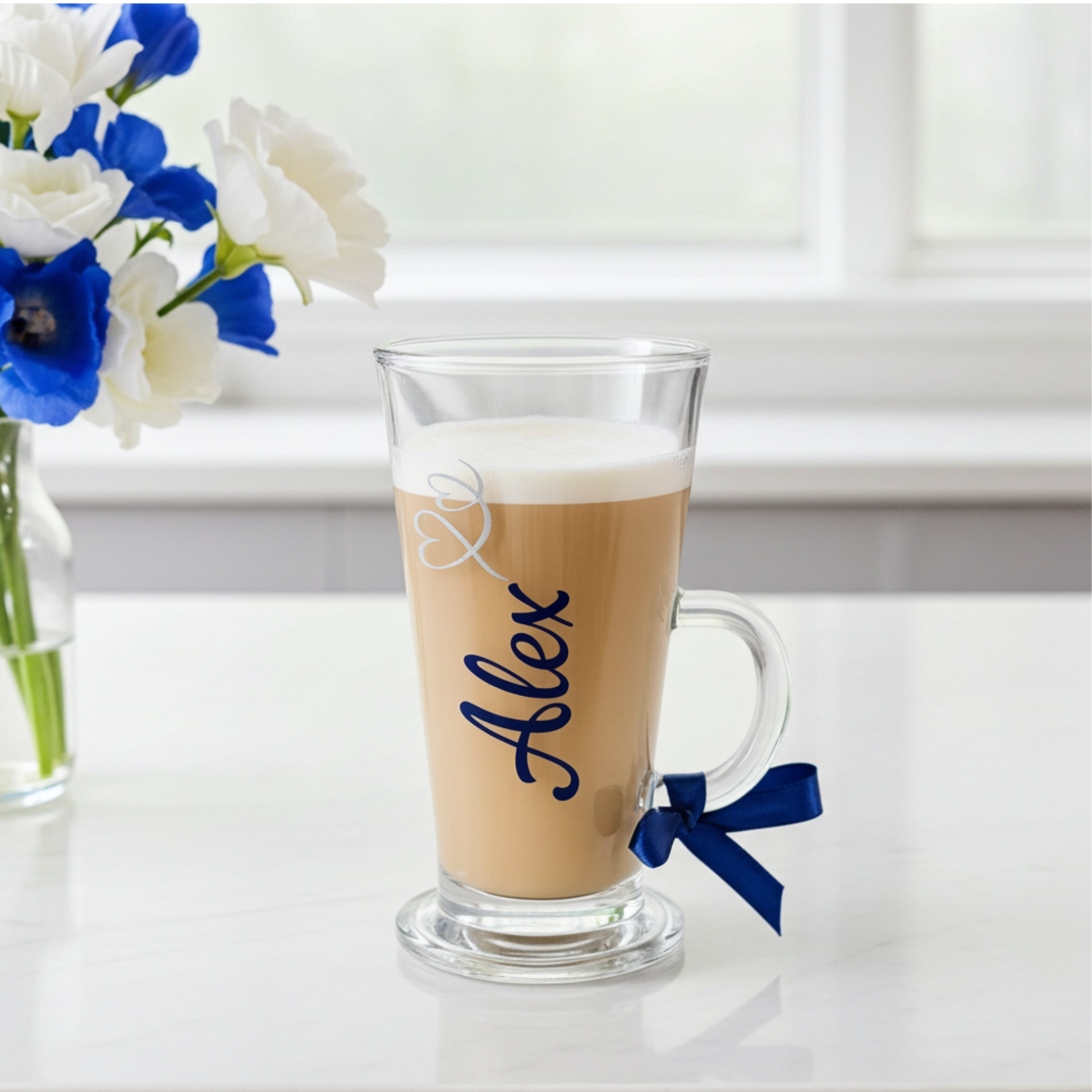 Personalised latte glass mug with the text 'Alex' on it in a cobalt blue vinyl. Features a silver glitter heart swirl design and a cobalt blue satin ribbon tied around the handle. Sat on a white marble surface next to a vase of flowers.