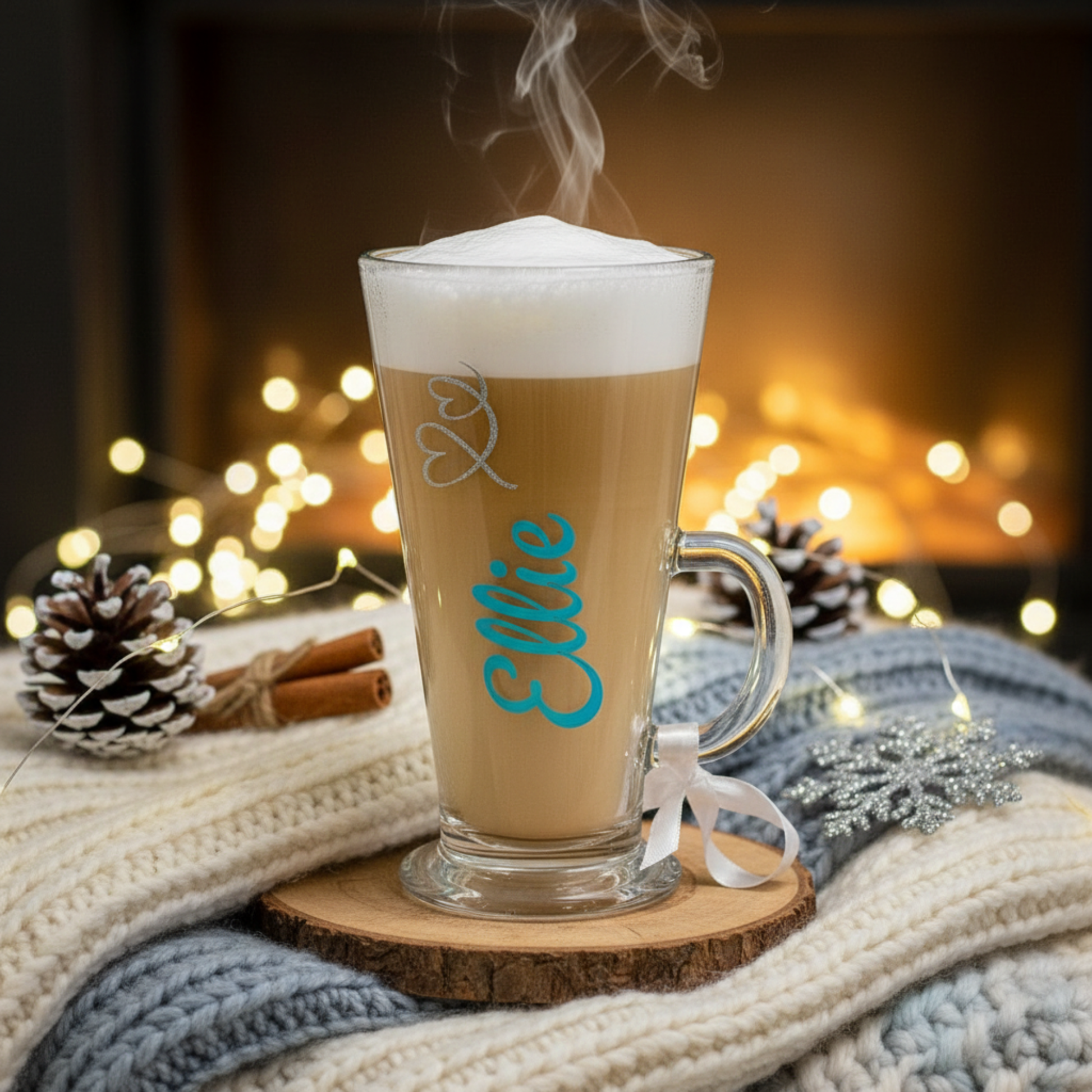 Steaming latte in a glass with 'Ellie' on a wooden coaster, surrounded by festive decorations.