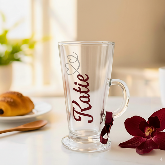 A clear latte glass. Has the name 'Katie.' On a surface next to a burgundy flower 