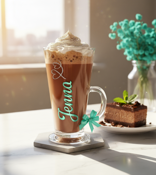 Glass of hot chocolate with whipped cream, a small cake, and a vase of flowers on a table.