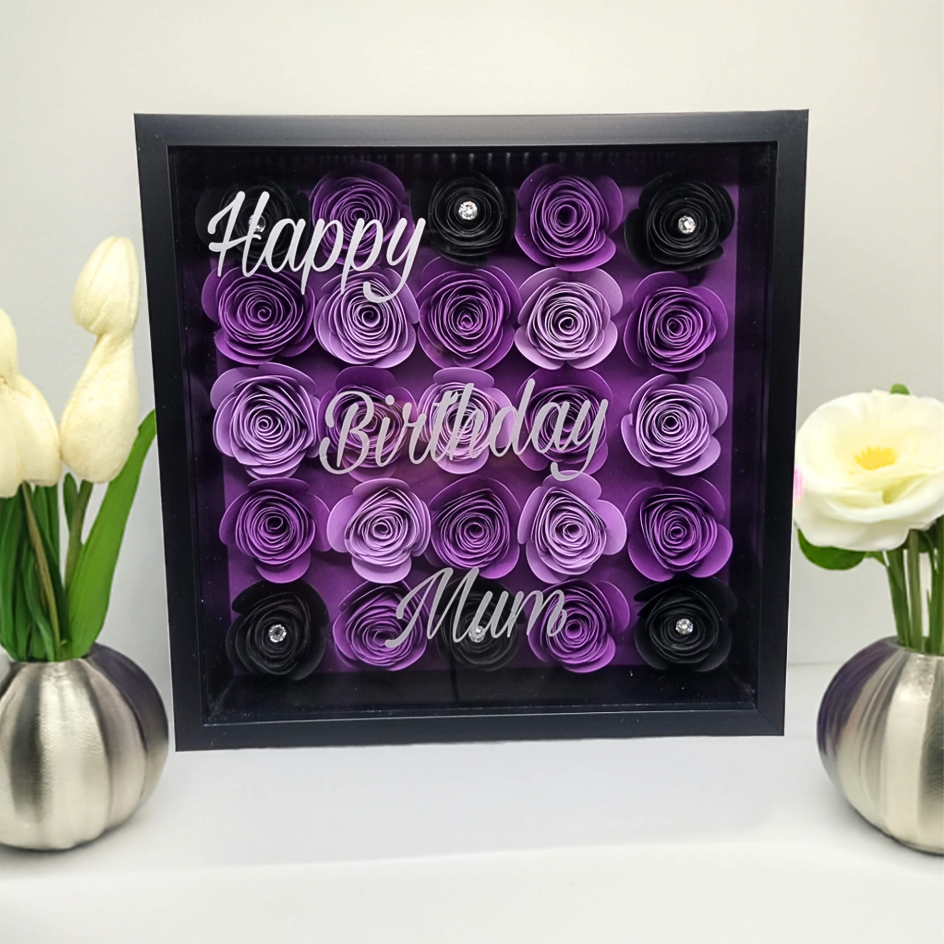 Shadow box with purple & black flowers behind a glass fronted frame with the words 'Happy Birthday Mum.' Sits on a shelf next to vase of flowers 