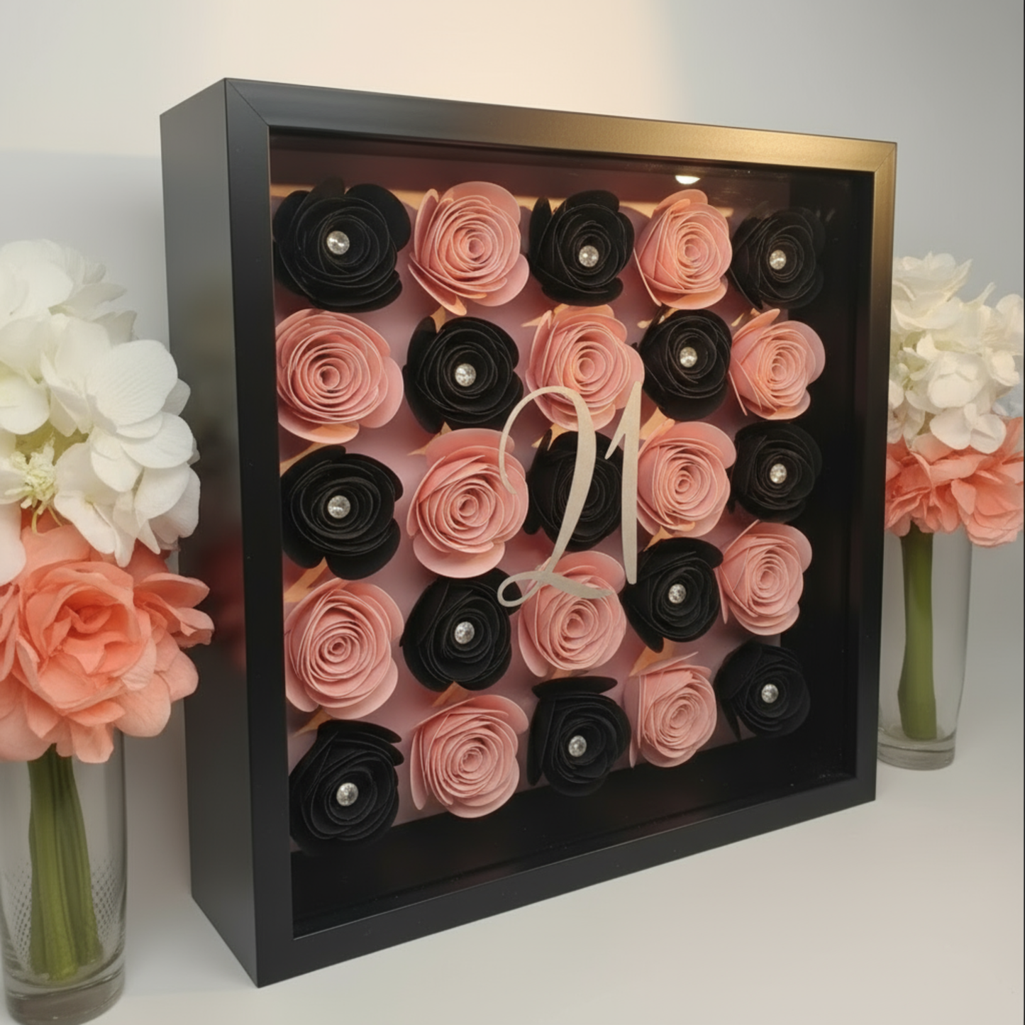 Personalised pink 3D shadow box with the age '21' on a glass front in a black square frame next to vases of flowers each side.