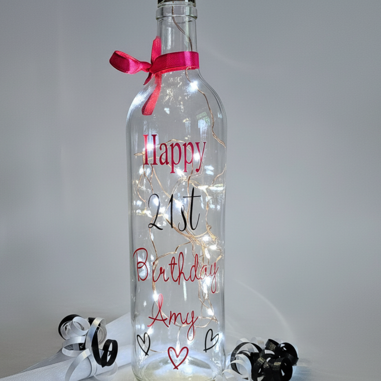 Personalised LED light up glass bottle with 'Happy Birthday Amy' in a pink vinyl text & age '21st' in black vinyl text next to an organza gift bag & satin curling ribbons.