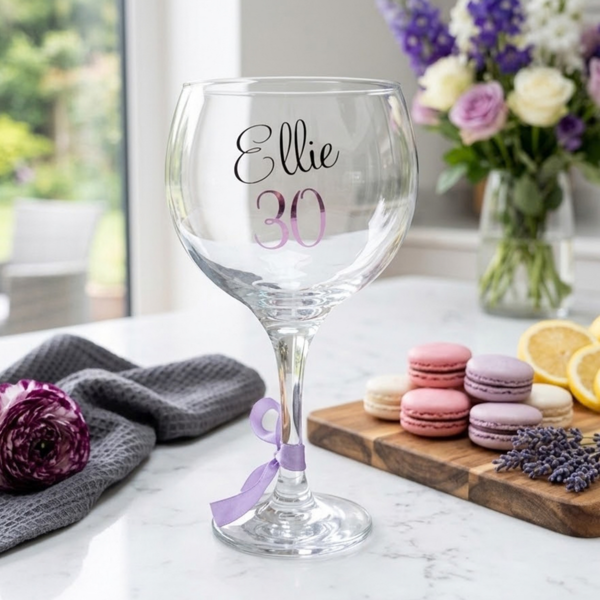 Personalised balloon gin glass with the name 'Ellie' in a flowing script font designed from a glossy black vinyl. This cocktail goblet has age '30' in a pink purple chrome holographic vinyl and a lavender satin ribbon around its stem. Styled on a white surface.