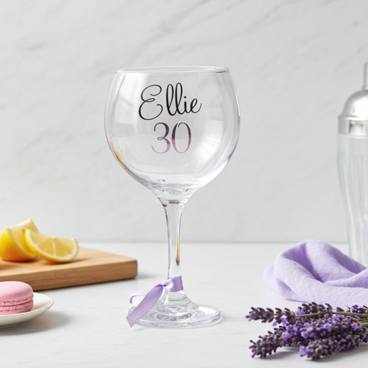 Personalised gin glass with the name 'Ellie' in a flowing script font designed from a glossy black vinyl. This cocktail goblet has the age '30' in a pink purple chrome holographic vinyl and a matching satin ribbon around its stem. Placed on a white surface next to lavender sprigs & choppiing board of sliced lemons.