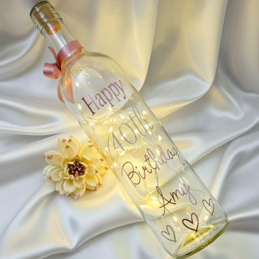 Personalised 40th birthday LED glass bottle lamp. Has the text 'Happy 40th Birthday Amy' in rose gold & silver vinyl. Features a small hearts design & a rose gold satin ribbon. Styled on a satin drape next to a flower.