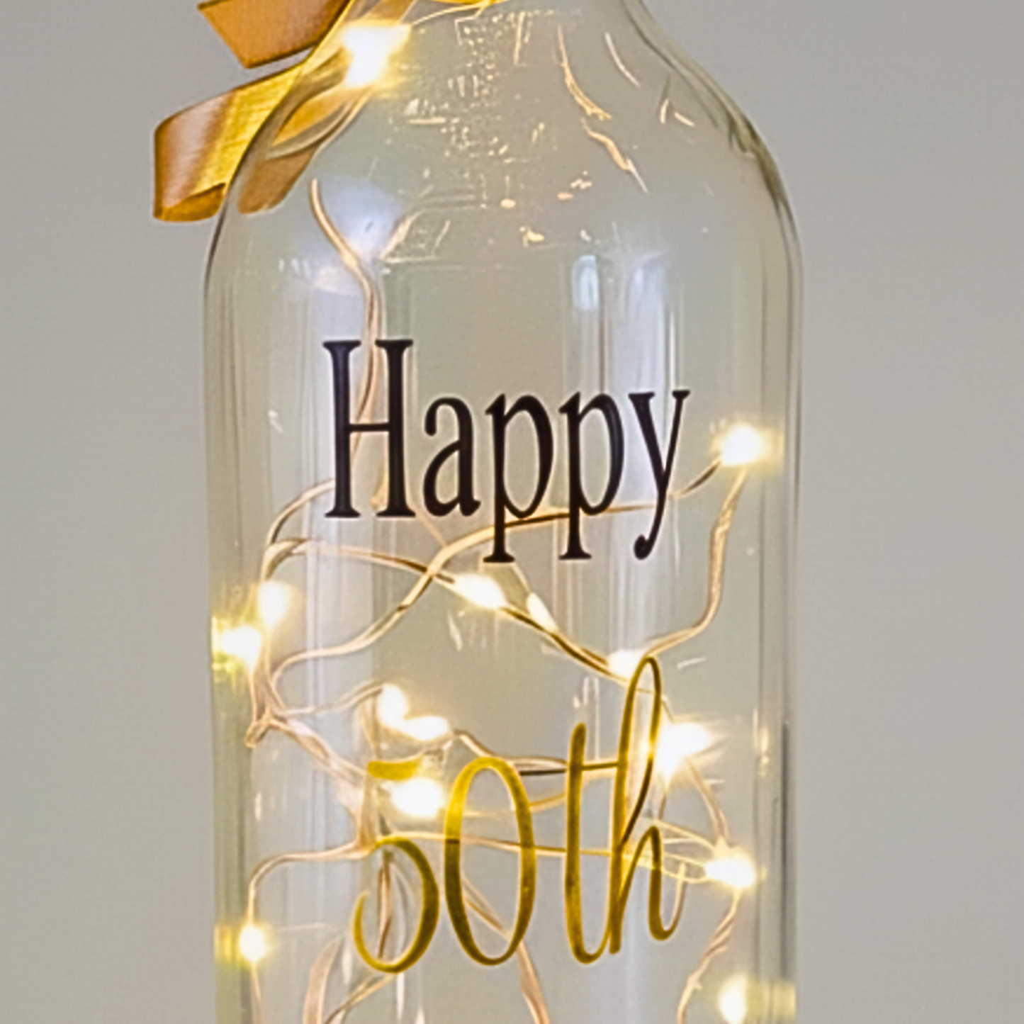 A clear glass bottle, displays 'Happy' in black vinyl and 50th in gold vinyl with a satin ribbon & LED lights 