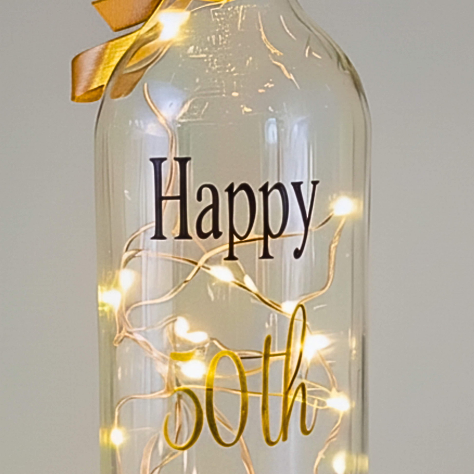 A clear glass bottle, displays 'Happy' in black vinyl and 50th in gold vinyl with a satin ribbon & LED lights 