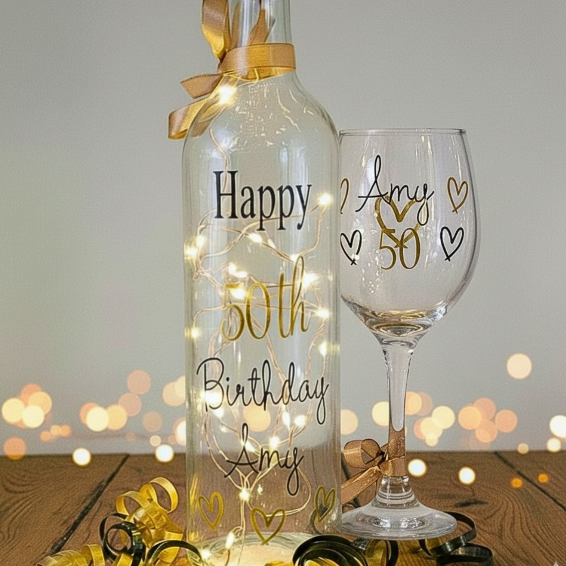 Personalised 50th birthday LED light up bottle and glass set with gold & black vinyl text. Set against a blurred fairy light background.