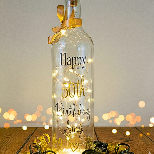 Personalised 50th LED light up glass bottle with 'Happy 50th Birthday Amy' text in gold & black vinyl text on a wooden surface with satin curling ribbons around.
