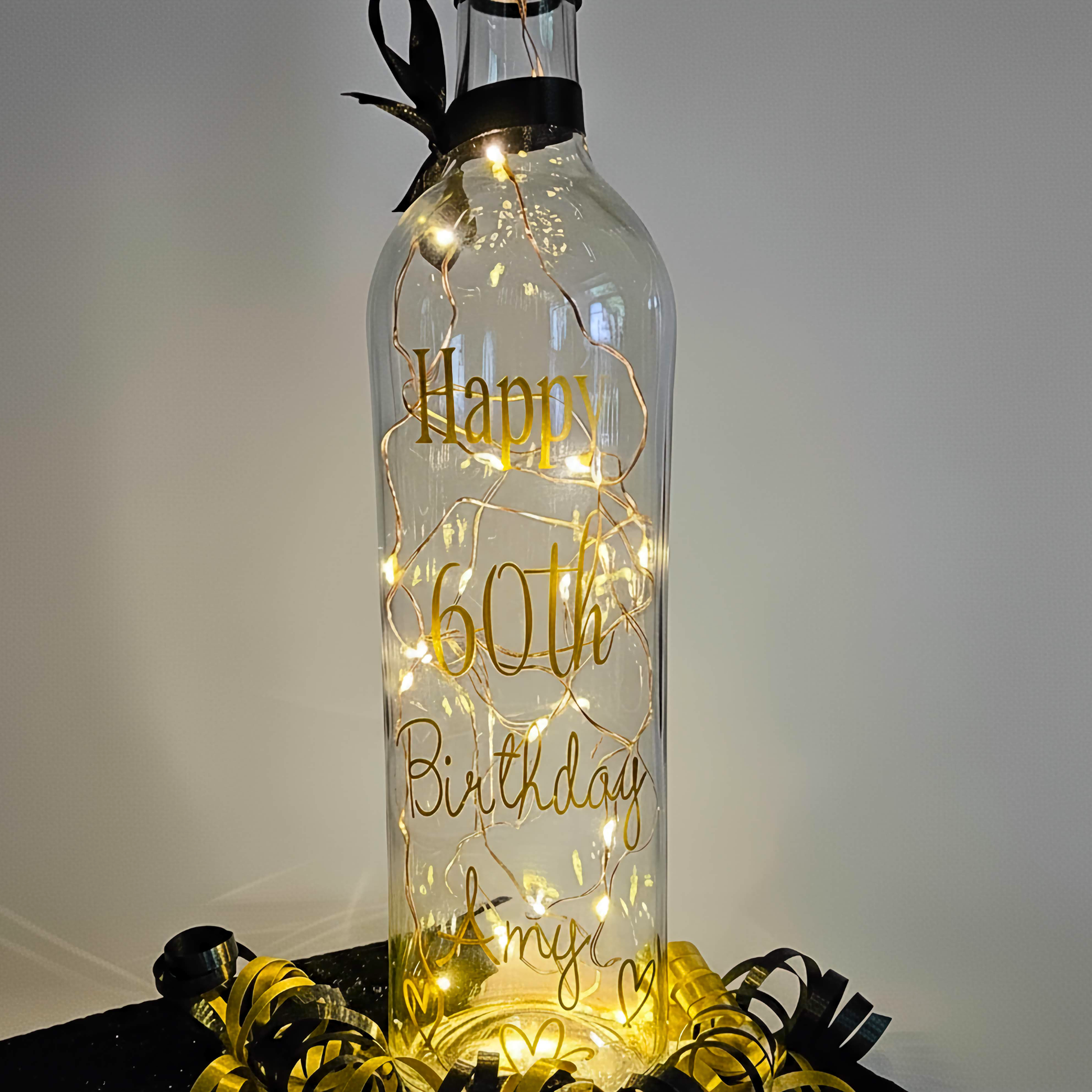 Personalised LED light up glass bottle with gold vinyl text 'Happy 60th Birthday Amy' next to an organza gift bag with satin curling ribbons around.