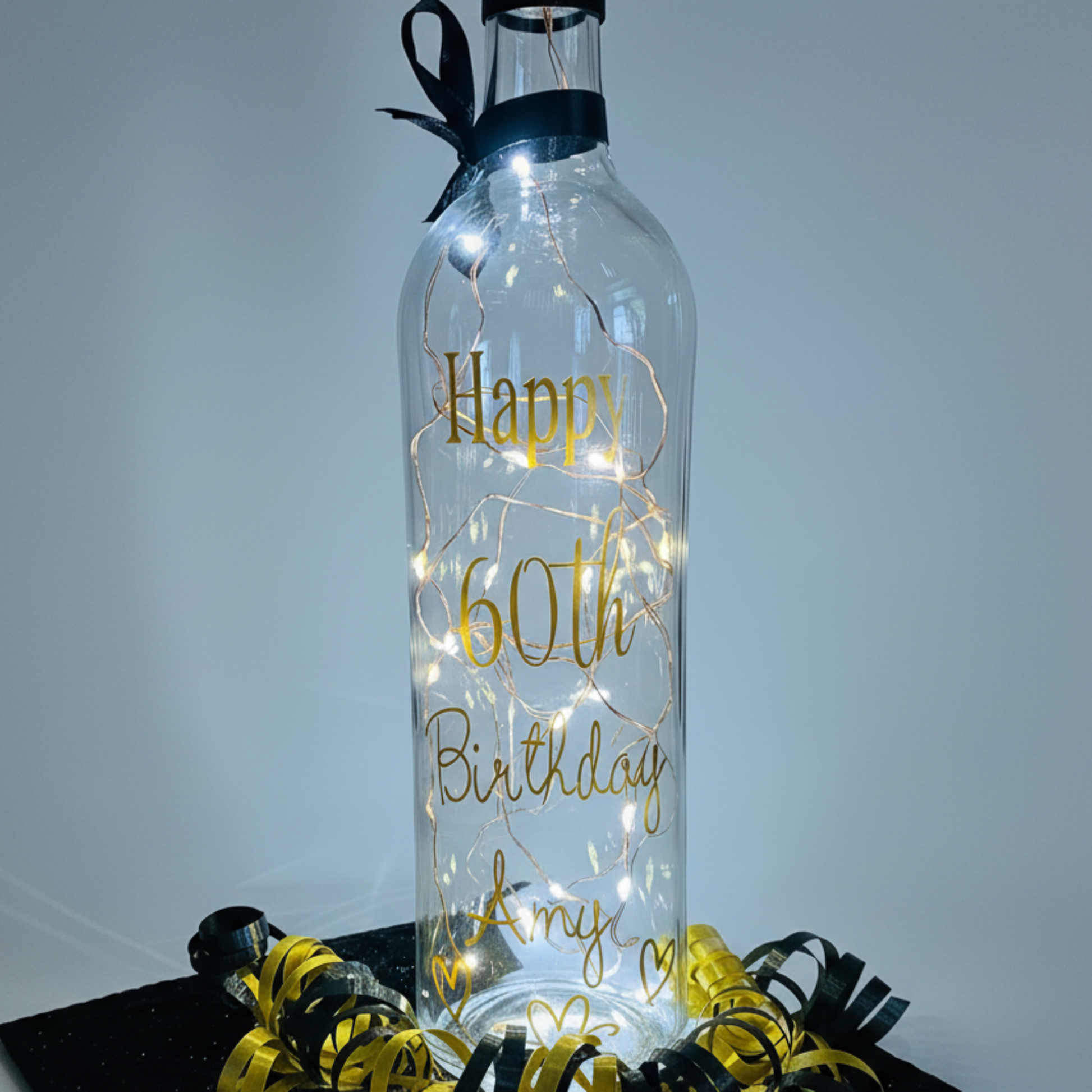 Personalised LED light up glass bottle with gold vinyl text 'Happy 60th Birthday Amy' next to an organza gift bag with satin curling ribbons around.