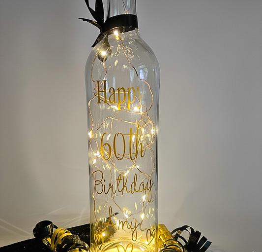 A clear bottle with lights inside. Wording 'Happy 60th Birthday Amy' & hearts in gold with a black ribbon to the neck. Has curling ribbons around.