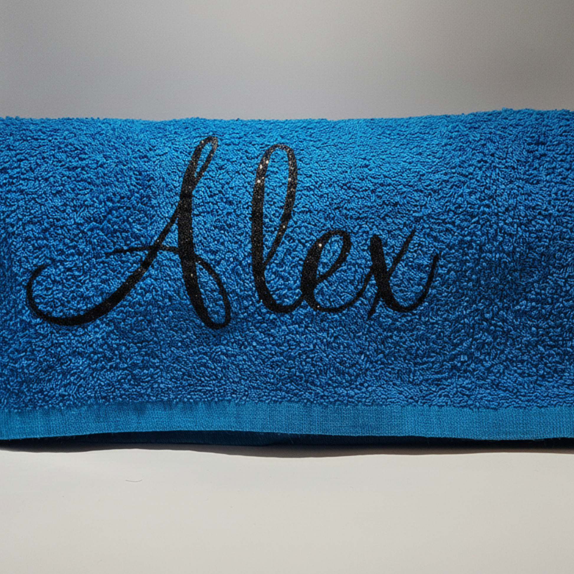 Personalised royal blue beach towel with the name 'Alex' in a black glitter vinyl on a neutral background