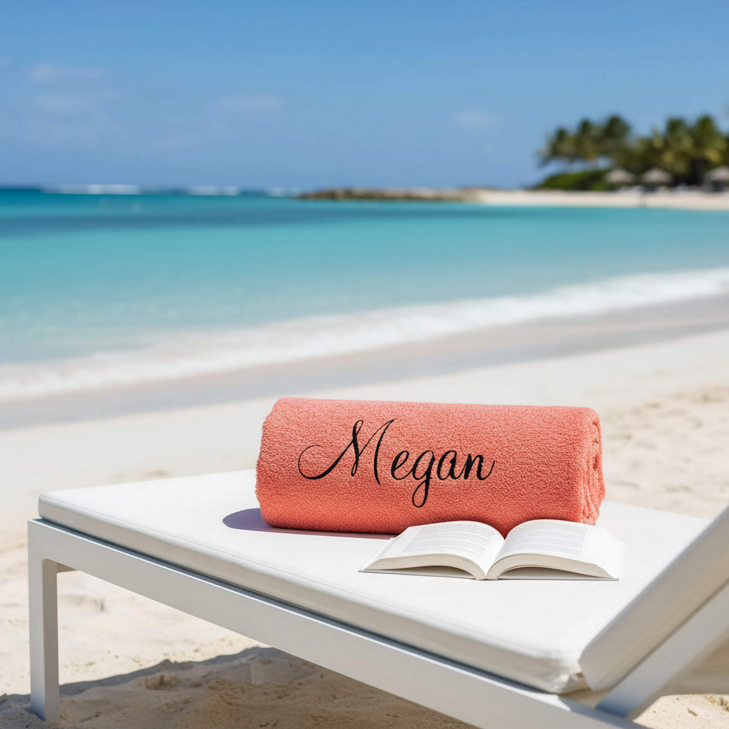 A personalised salmon pink colour beach towel with the name 'Megan' on it on a white sunbed on a beach by the sea with palm tree background.