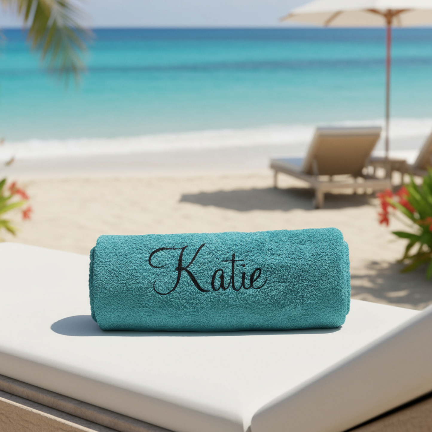 A personalised turquoise beach towel with the name 'Katie' on it in a black glitter vinyl text laid on a sunbed by the beach & ocean