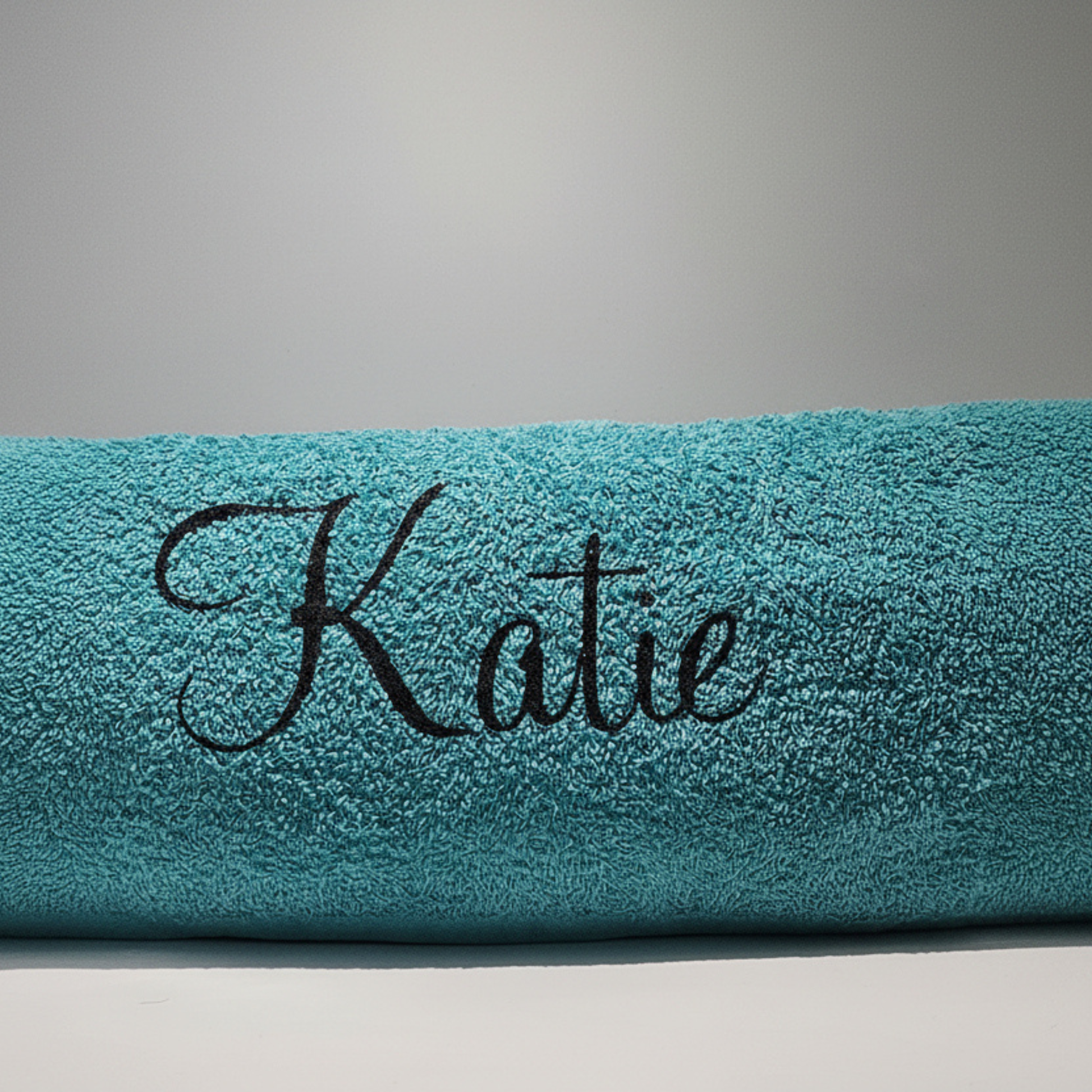 Personalised beach towel in turquoise with the name 'Katie' on it in a black glitter vinyl text on a plain background.