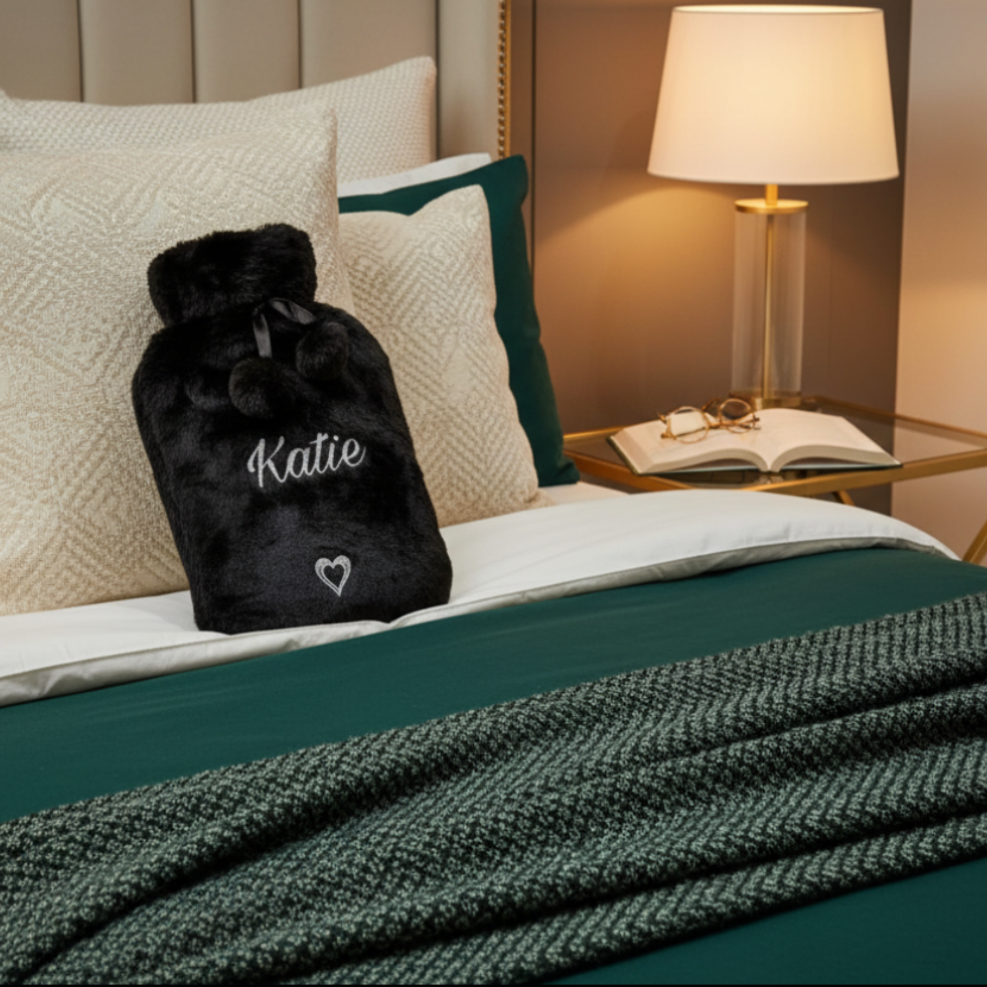 Personalised faux fur hot water bottle in black with the name 'Katie' text & heart feature in silver glitter vinyl. Laid on a bed up against cushions. Next to a bedside table with lamp & book.