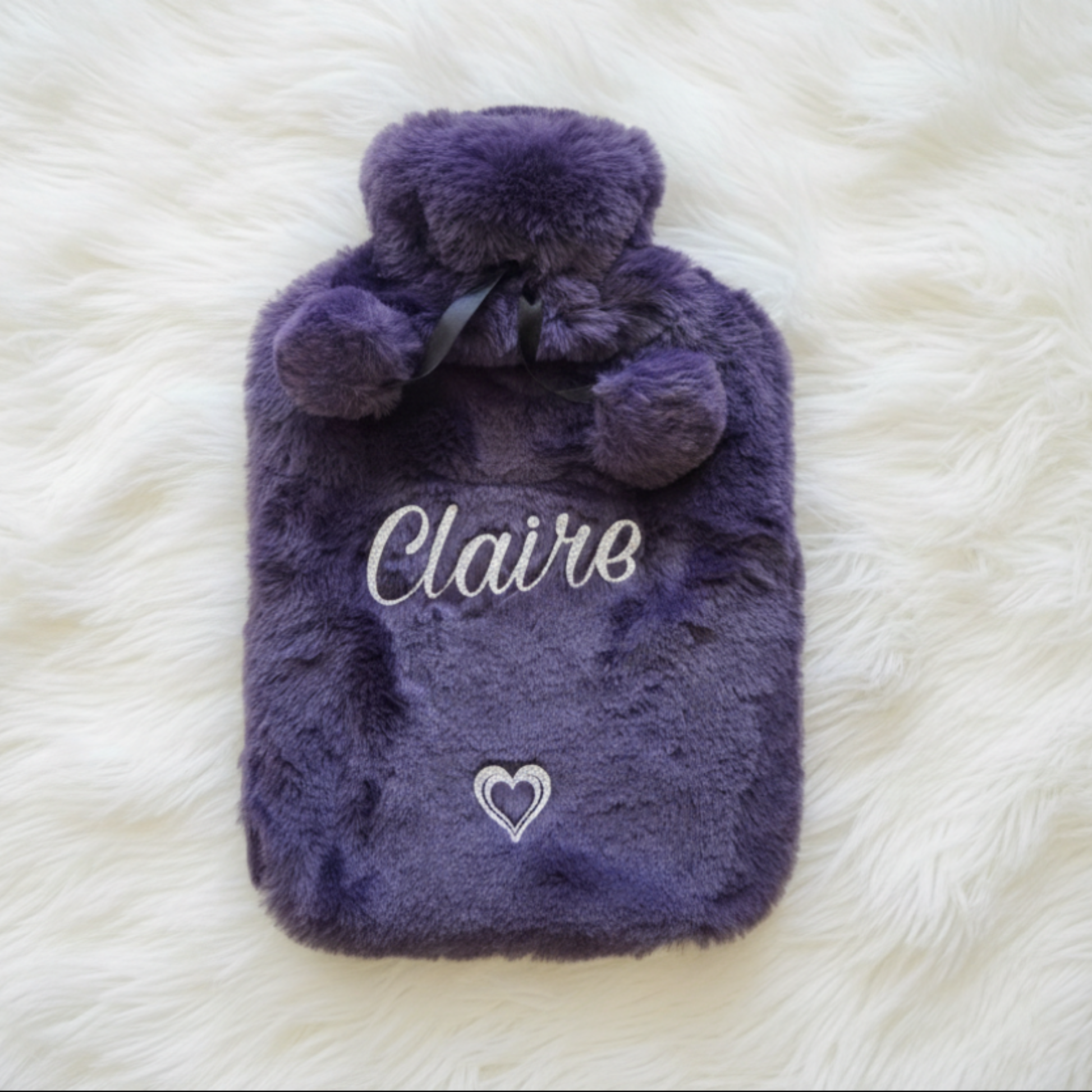 Personalised faux fur hot water bottle with pom poms in purple with the name 'Claire' in a silver glitter vinyl text with heart design laid on a fluffy white blanket.