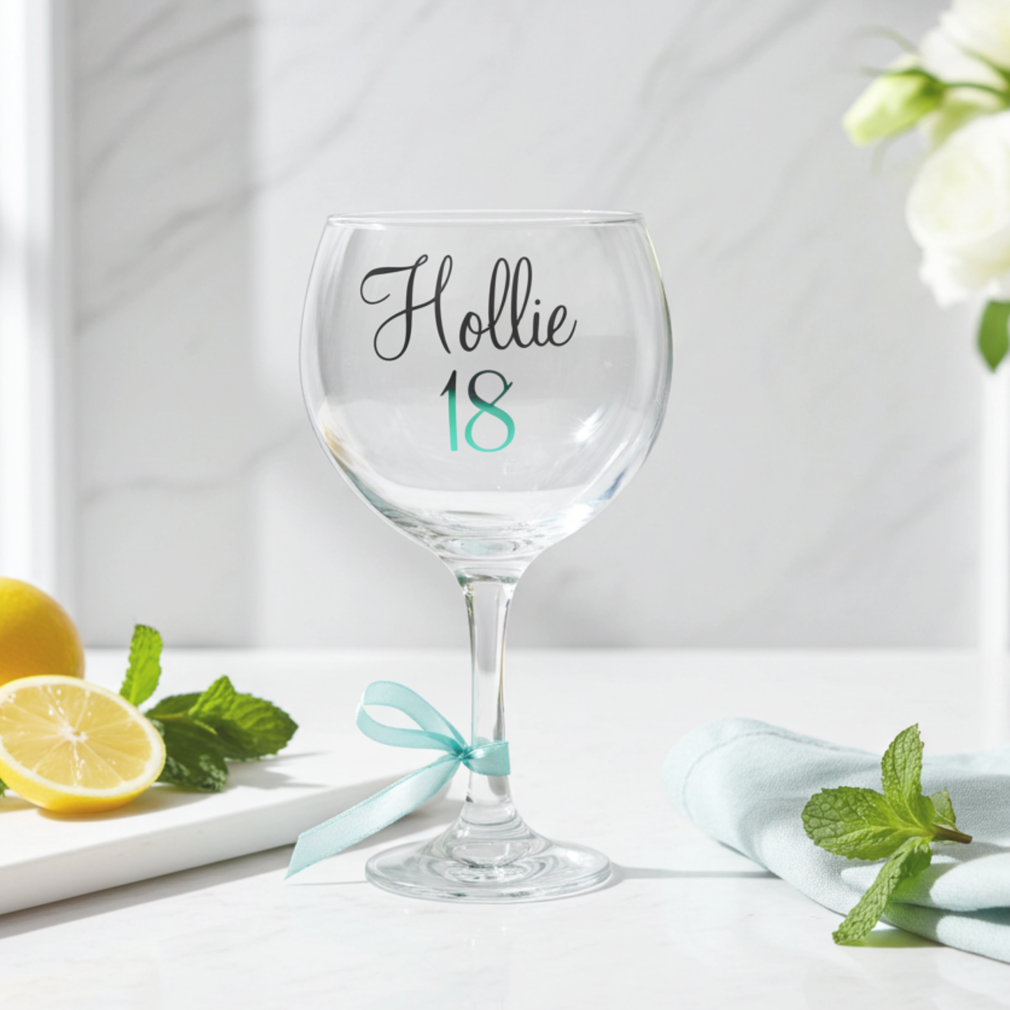 Personalised balloon gin glass with the name 'Hollie' in a flowing script font from a glossy black vinyl. This stemmed cocktail goblet features age '18' in a mint chrome holographic vinyl & a mint satin ribbon tied around its stem. Placed on a white marble surface next to a chopping board of lemon slices & mint sprigs. 