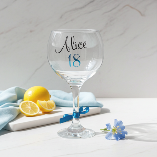 Personalised gin glass with the name 'Alice' in a flowing script font designed from a glossy black vinyl. This stemmed cocktail goblet features the age '18' in a peacock blue holographic chrome vinyl and a matching satin ribbon around its stem.  Placed on a white marble surface next to a chopping board of lemons slices, a blue napkin and small flower.