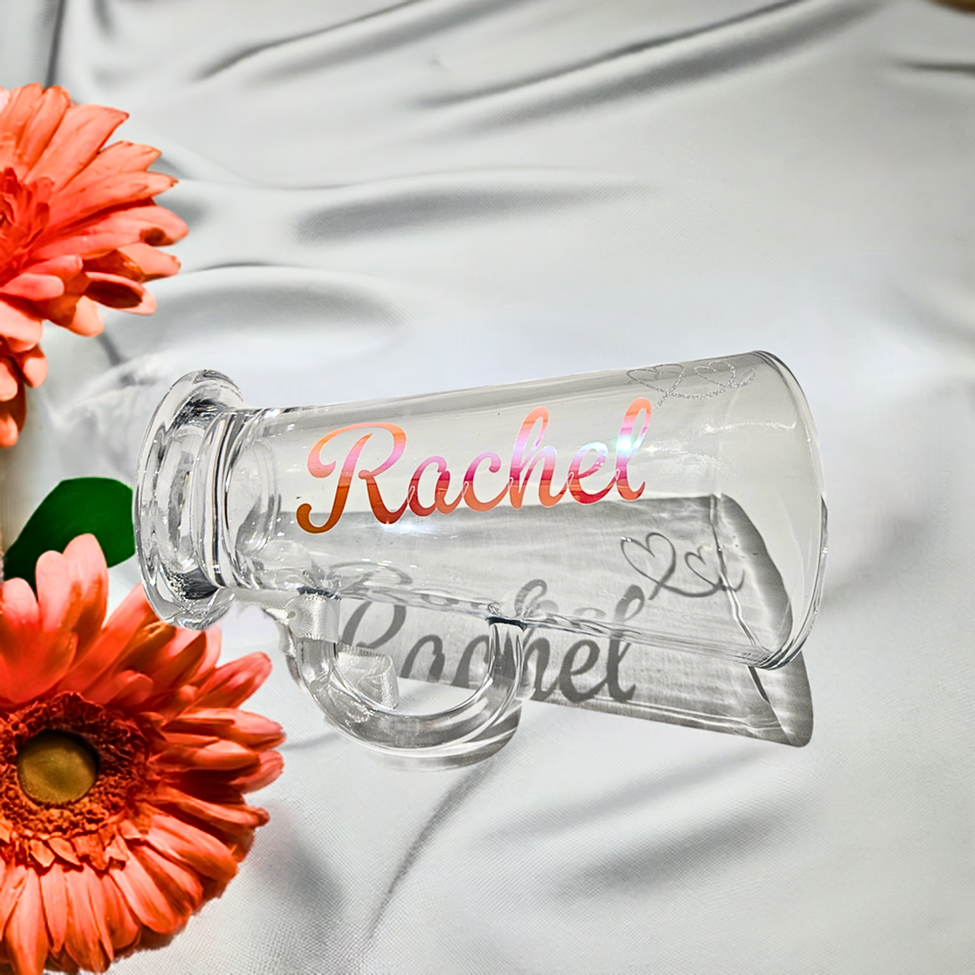 Clear glass mug with 'Rachel' in a holographic vinyl & silver glitter heart. Staged with orange flowers on a satin drape
