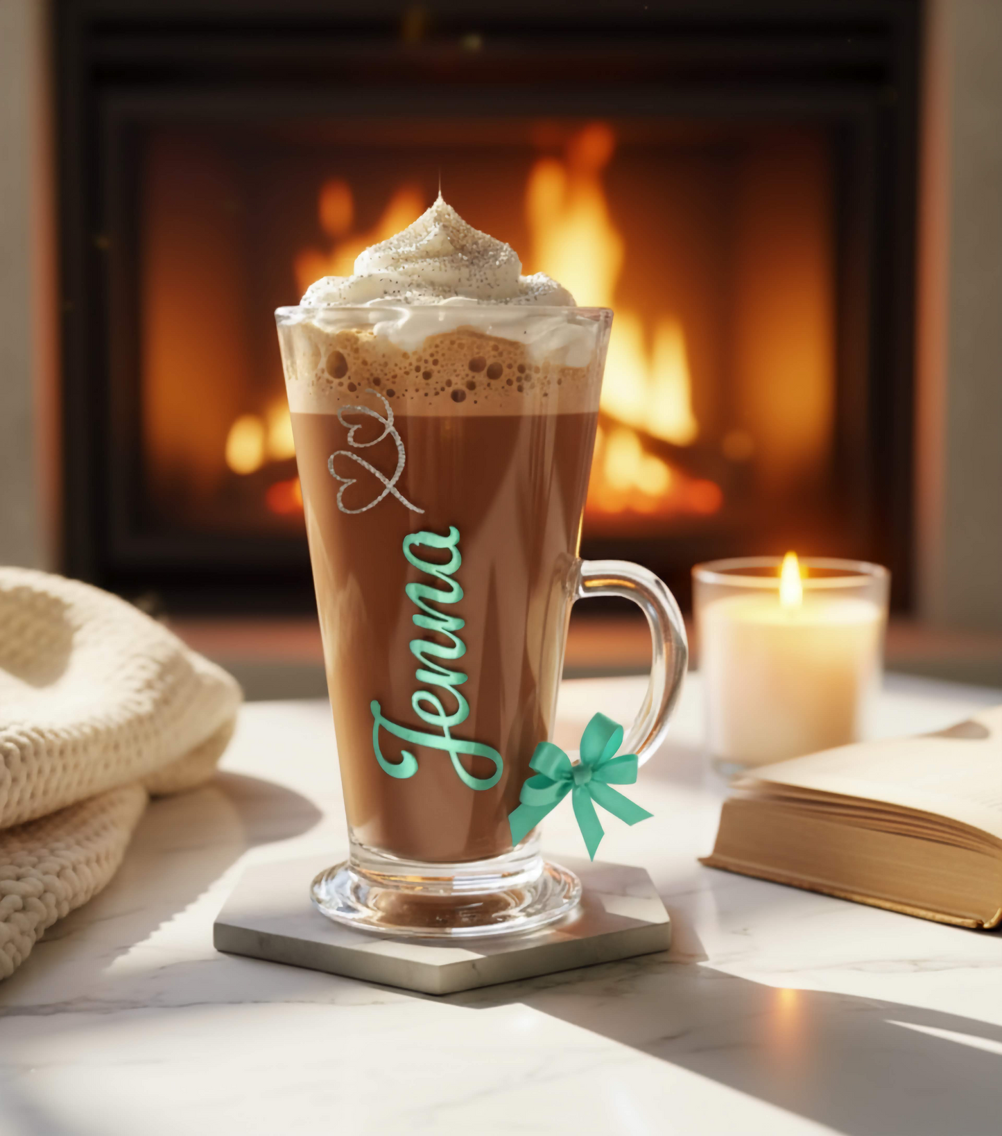 A clear glass mug filled with hot chocolate & whipped cream. Sat on a white surface next to a candle & book 