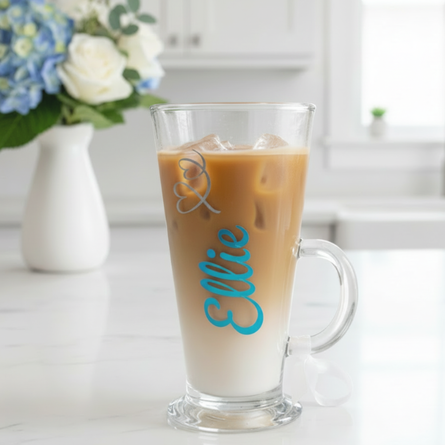 Personalised iced coffee, latte glass mug filled with iced coffee with the name 'Ellie' on it. Sat on a white kitchen top with a vase of blue & white flowers in the background.