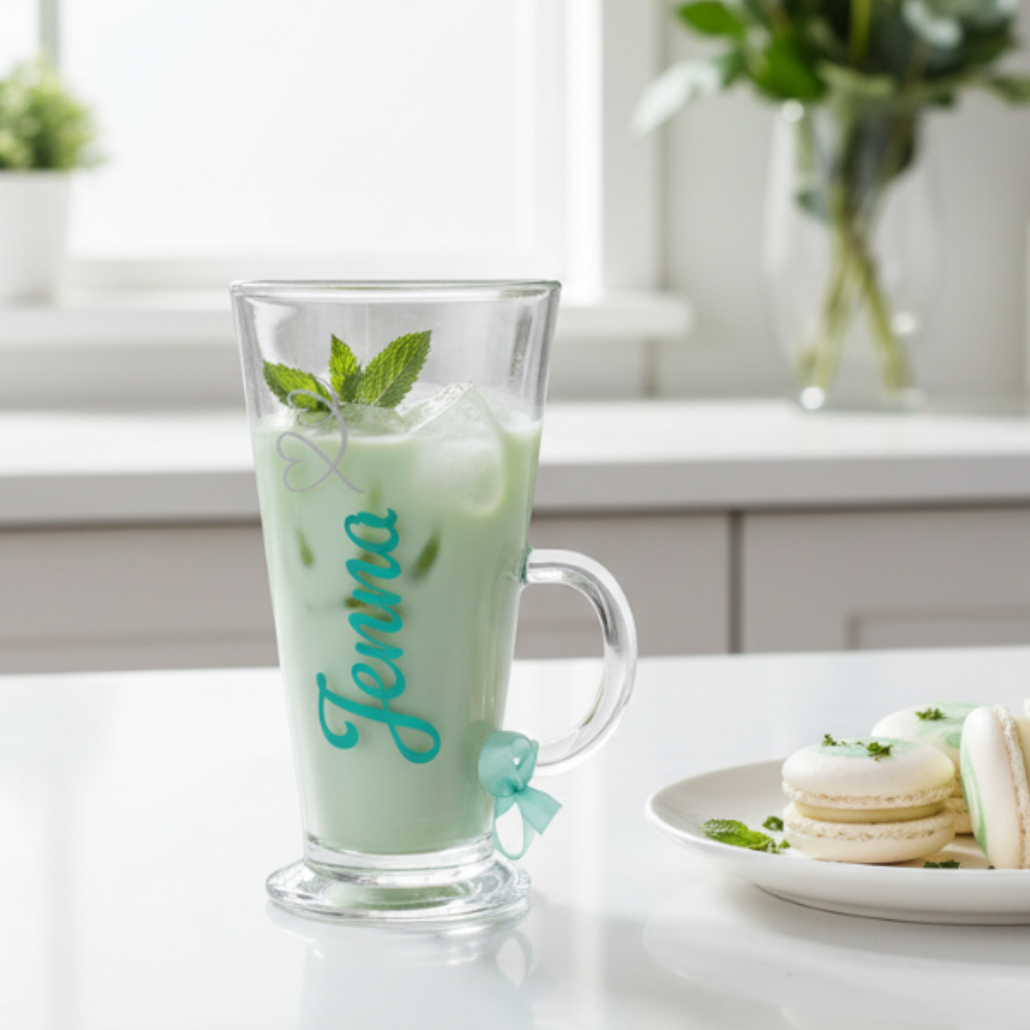 Personalised clear glass mug filled with an iced drink and mint leaves, with the name 'Jenna' on it sat on a white surface next to a plate of white & mint colour macaroons.