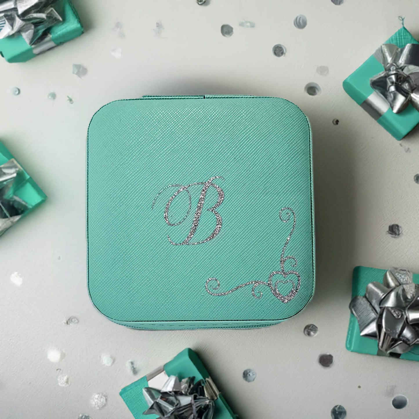 Turquoise jewellery box with a decorative 'B' on a light gray background with small gift boxes and ribbons.