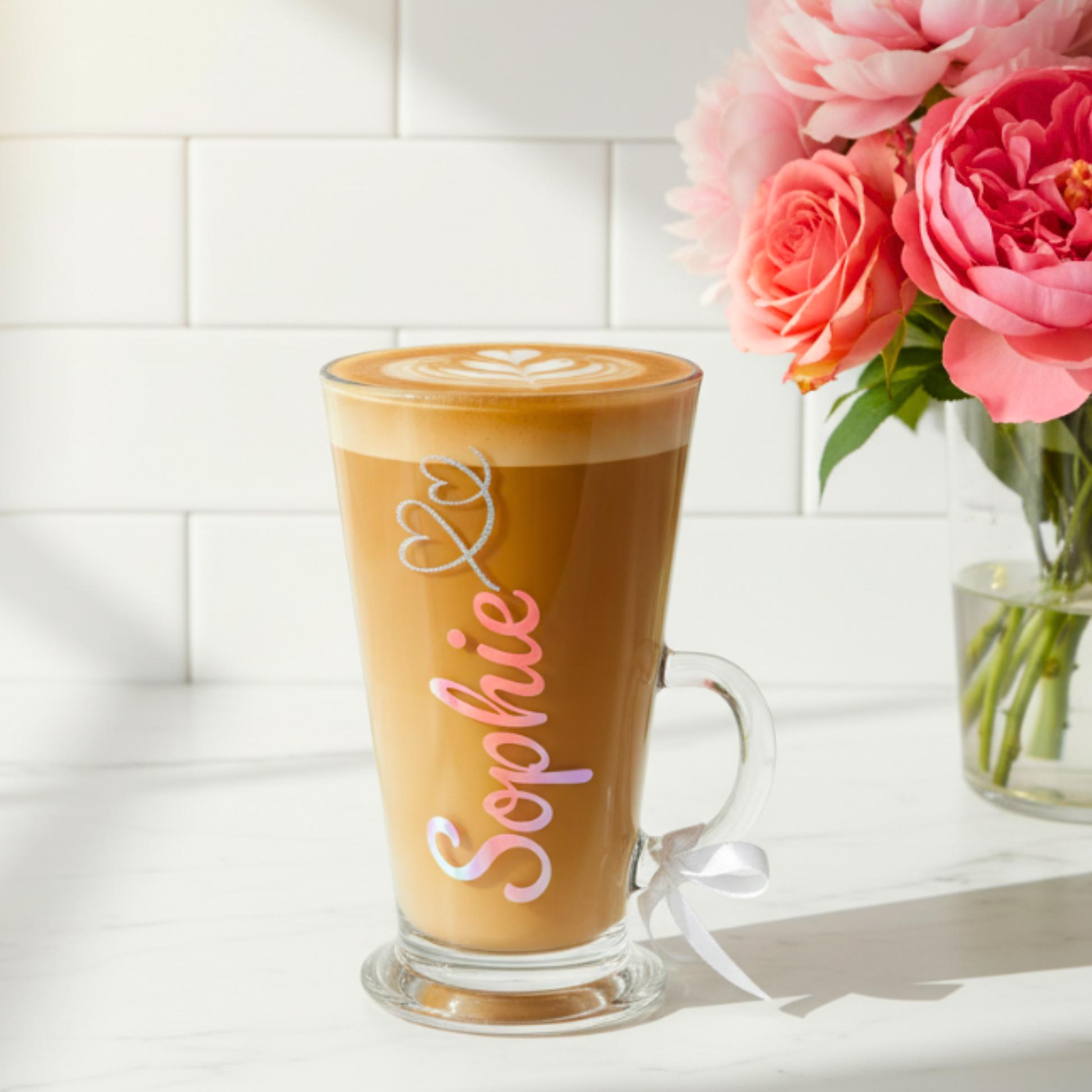 Personalised latte glass with the name 'Sophie' in a holographic pink coral vinyl. Features a silver glitter heart swirl  design with a white satin ribbon on the handle. Sits on a white surface next to a vase of pink flowers.