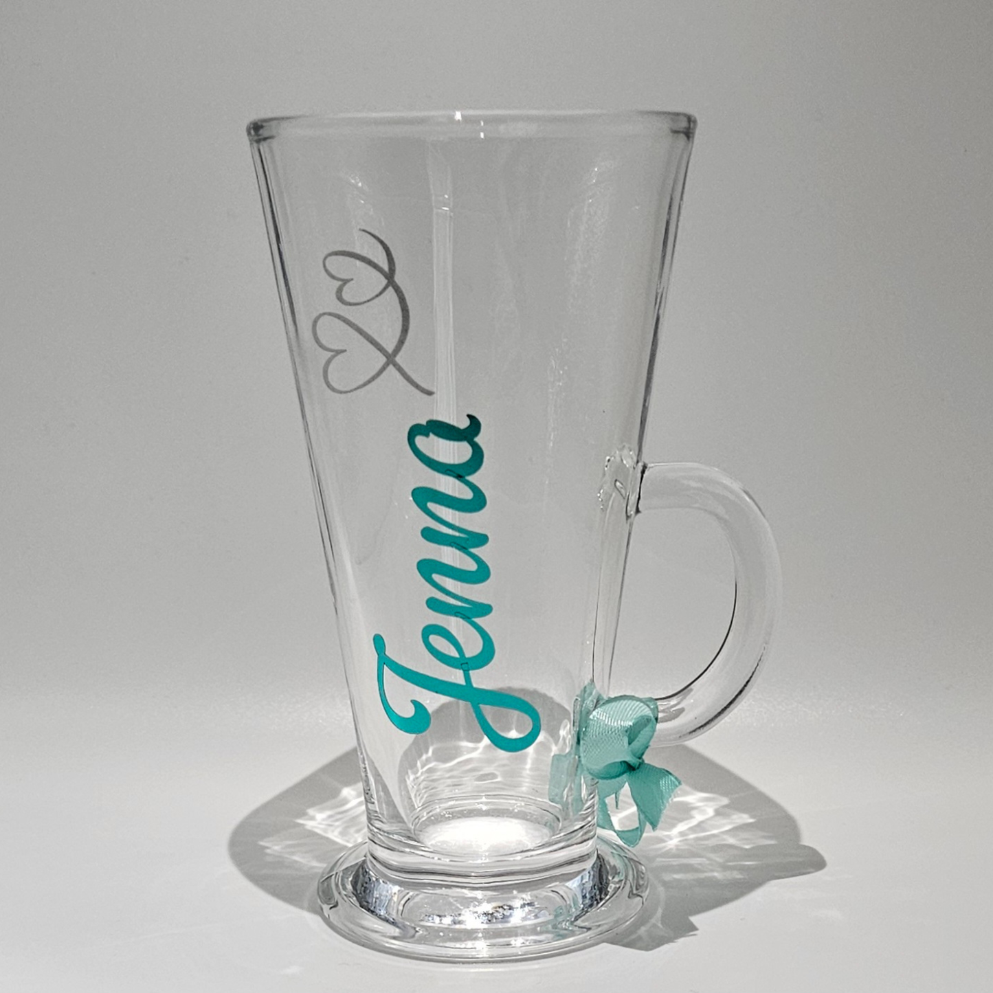 Personalised clear glass mug with the name 'Jenna' on it in a mint holographic vinyl with silver heart swirl on a light grey background