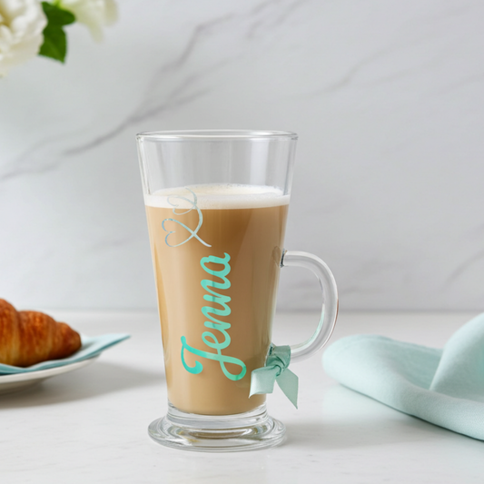 Personalised clear latte glass with the text 'Jenna' in a mint chrome holographic vinyl. This tapered coffee glass mug has a silver glitter hearts swirl design & a mint satin ribbon tied around its wide loop handle. Placed on a white surface next to a croissant & napkin.