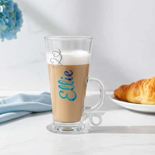 Personalised clear latte glass mug with the name 'Ellie' in a peacock blue chrome holographic vinyl. Has a frosted glitter heart swirl feature & a satin ribbon around its wide loop handle. On a white surface with a marble white background.