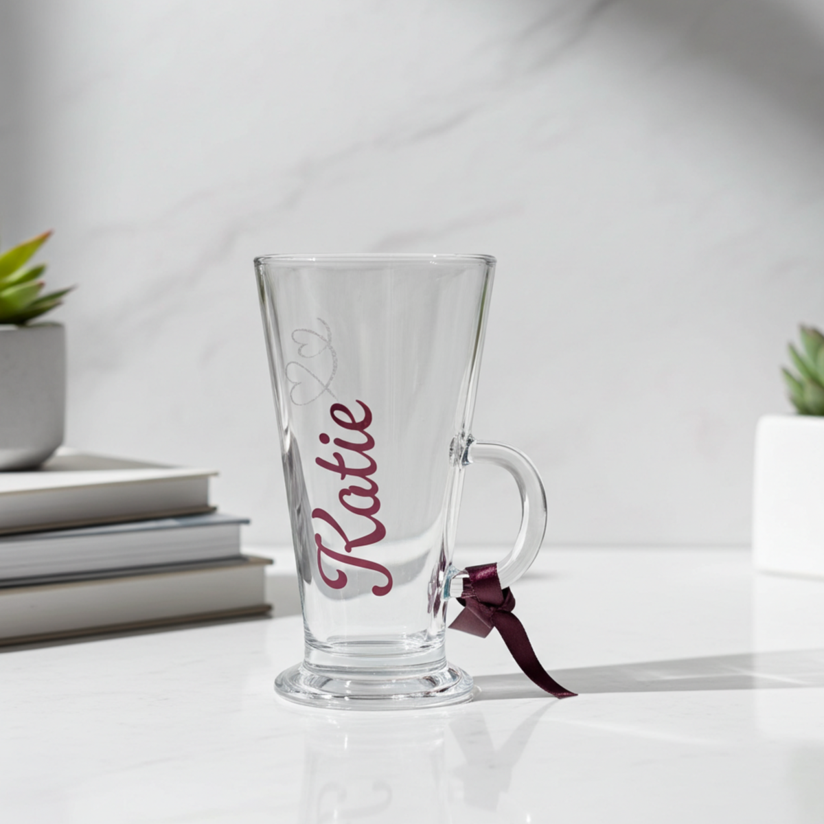 Personalised clear latte & hot chocolate glass with the text 'Katie' in a burgundy chrome holographic vinyl. This tapered coffee glass mug features a silver glitter hearts swirl design and a burgundy satin ribbon tied around its wide loop handle. Placed on a white counter top with a marble background.