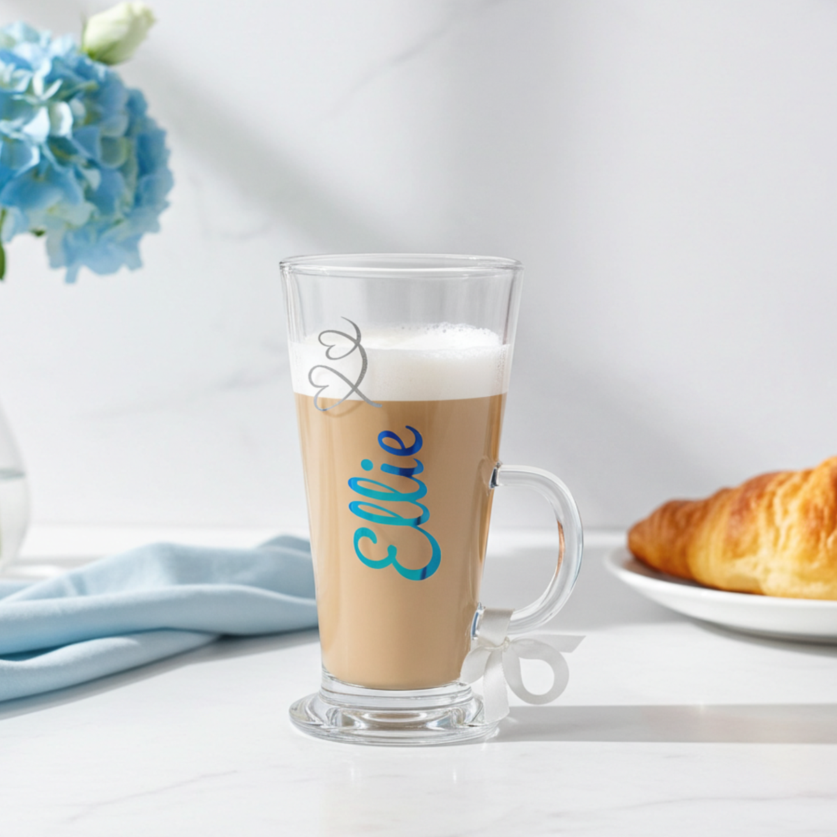 Personalised clear latte glass with the text 'Ellie' in a peacock blue chrome holographic vinyl. This tapered coffee glass mug has a silver glitter hearts swirl design & a white satin ribbon tied around its wide loop handle. Placed on a white marble surface next to a blue napkin, flowers and a croissant.