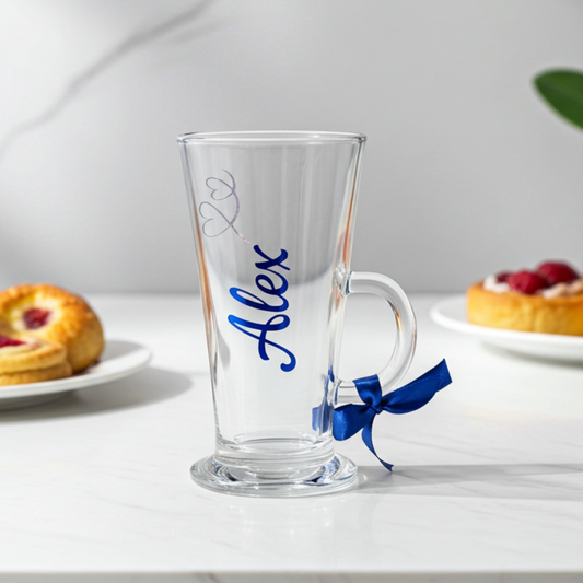 Personalised clear latte & hot chocolate glass with the text 'Alex' in a cobalt blue vinyl. This tapered coffee glass mug features a silver glitter hearts swirl design with a cobalt blue satin ribbon tied around its wide loop handle. Placed on a marble surface next to a jam pastry.