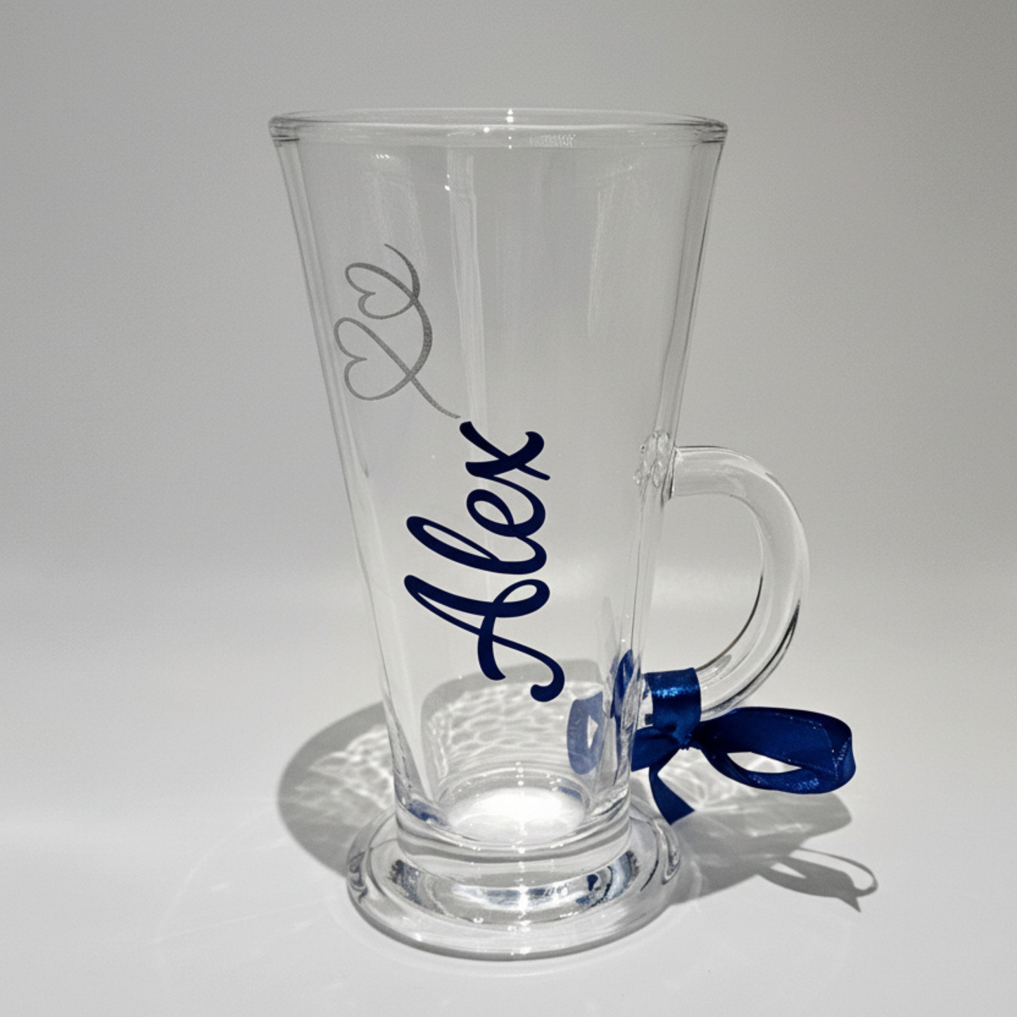 Personalised clear latte glass mug with the name 'Alex' in a cobalt blue chrome vinyl. Featuring a blue ribbon on a white background.